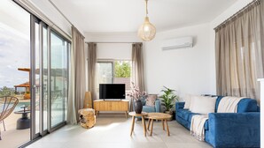 A spacious living room with a large sliding glass door leading to a balcony, a blue sofa, a wooden coffee table, and a television.