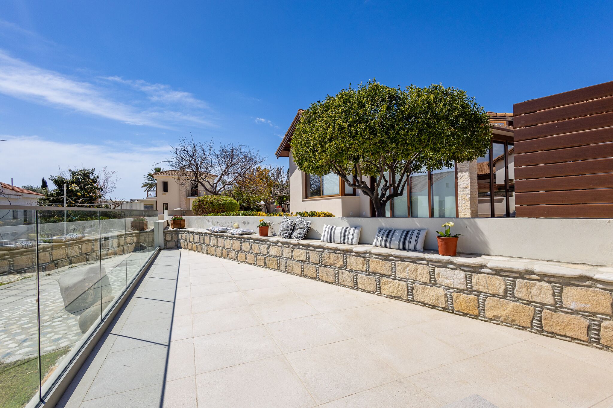 A modern outdoor patio area with a stone wall, seating with cushions, and a large tree providing shade.