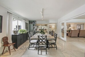A spacious dining area with a long table, surrounded by chairs, and a kitchen area with cabinets and a countertop. - Private Family Villa at Casa de Campo (La Romana)