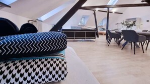 A modern loft-style room with a sloped ceiling, a piano, a dining area with chairs, and a large window. - Alpha Apartments: Grand Loft for 8, Roof Terrace (Krefeld)