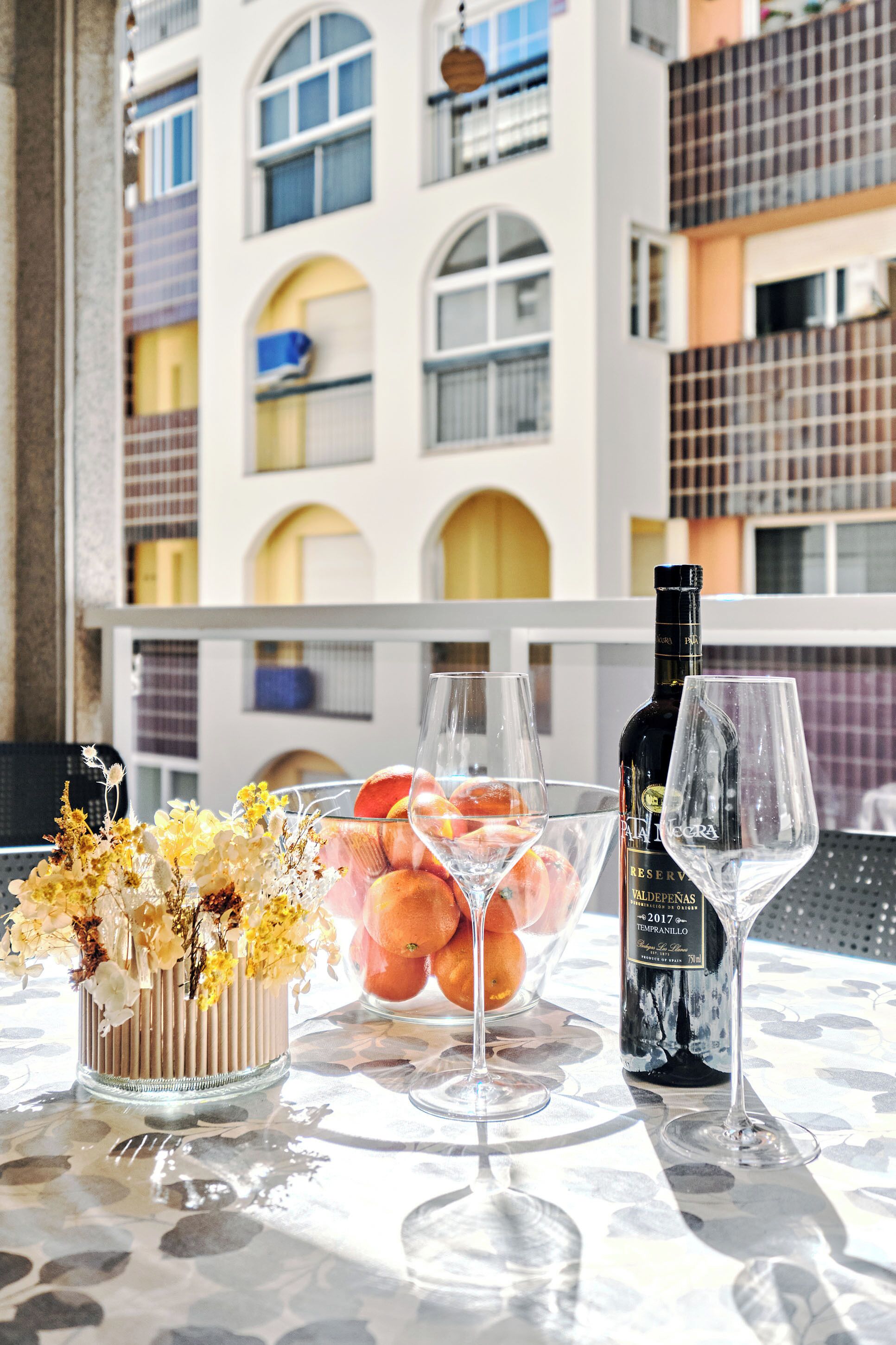 A table with a bottle of wine, two glasses, and a bowl of fruit on a balcony with a building in the background.