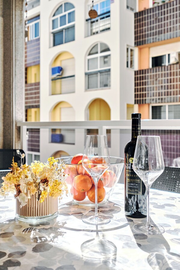 A table with a bottle of wine, two glasses, and a bowl of fruit on a balcony with a building in the background.