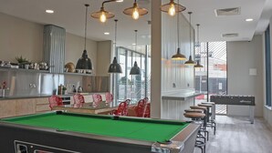 A pool table with a green felt surface, a billiards table, and a foosball table in a modern indoor setting with pendant lighting and a view of the outside.