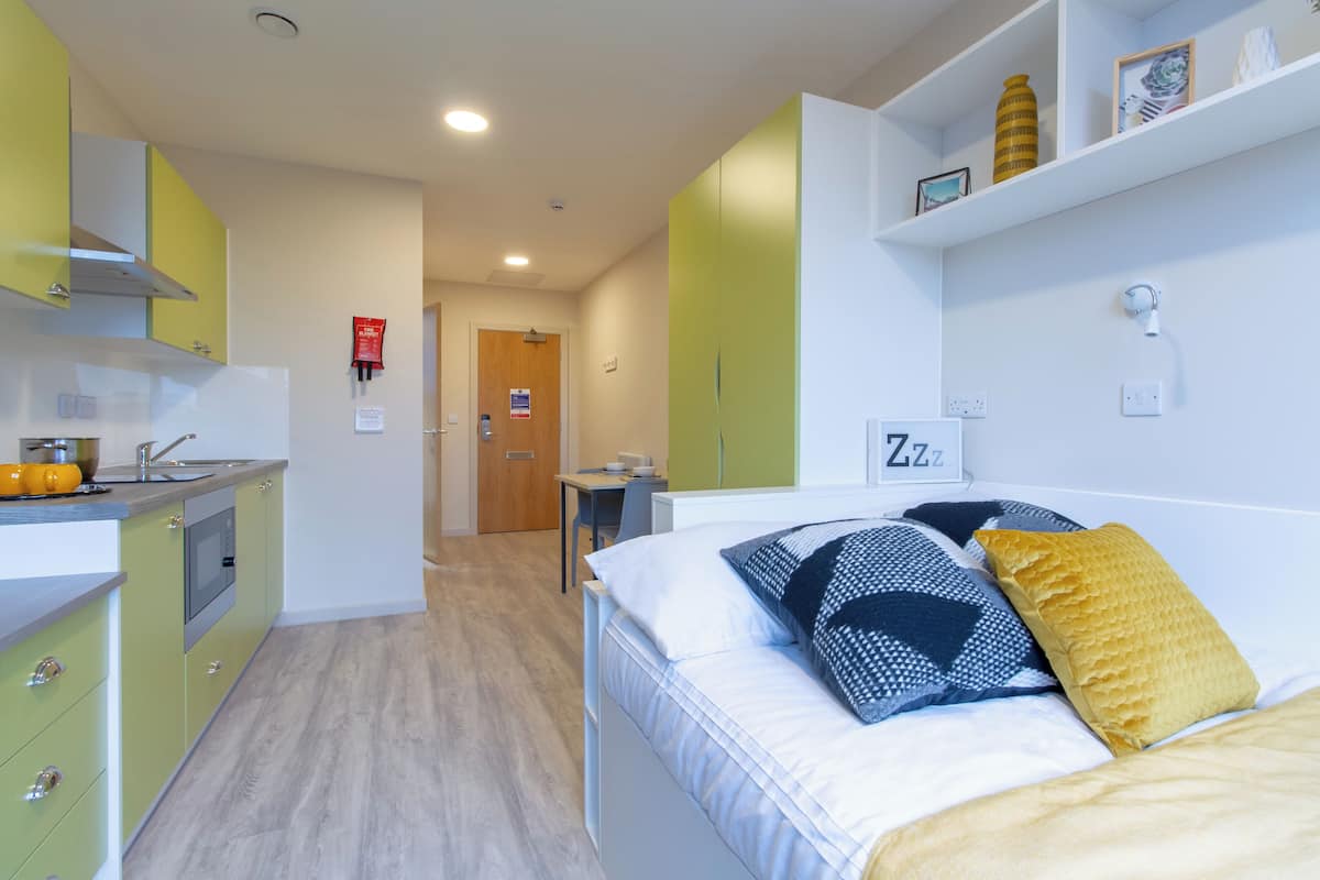A modern bedroom with a bed, bedside table, and a nightstand. There is a kitchen area with cabinets and a microwave.