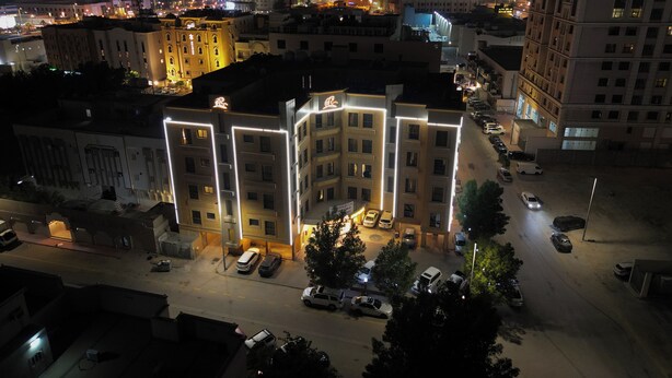 A multi-story building with illuminated outlines, surrounded by parked cars and other buildings.
