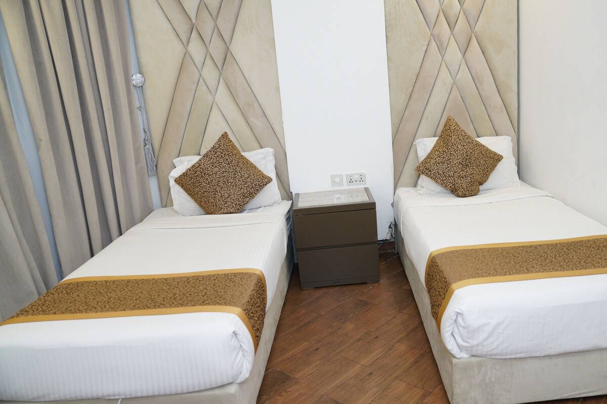 Une chambre d’hôtel avec deux lits simples, chacun recouvert d’une couverture à motifs et agrémenté d’oreillers assortis. Une petite table de chevet se trouve entre les lits, avec une lampe et une prise électrique. La chambre a un plancher en bois et un mur blanc uni.