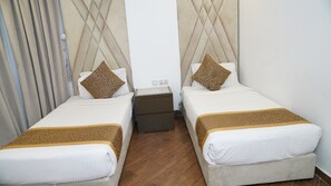 Et hotelværelse med to enkeltsenge, hver med et mønstret tæppe og matchende puder. Der er et lille natbord imellem sengene med en lampe og en stikkontakt. Værelset har trægulv og en ensfarvet, hvid væg.