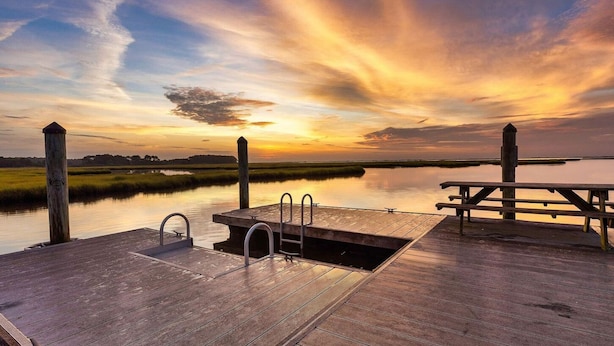 Une jetée en bois avec un banc et une petite piscine, d’où l’on peut admirer un plan d’eau paisible au coucher du soleil.