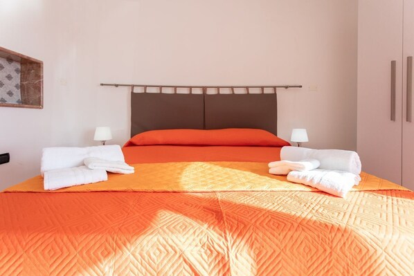 Ein ordentlich bezogenes Bett mit orangefarbener Tagesdecke und Kissen, zwei Nachttischlampen und ein Kopfteil mit Holzpaneelen.