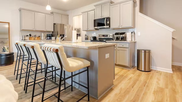 A modern kitchen with white cabinets, a central island with bar stools, stainless steel appliances, and a trash can.