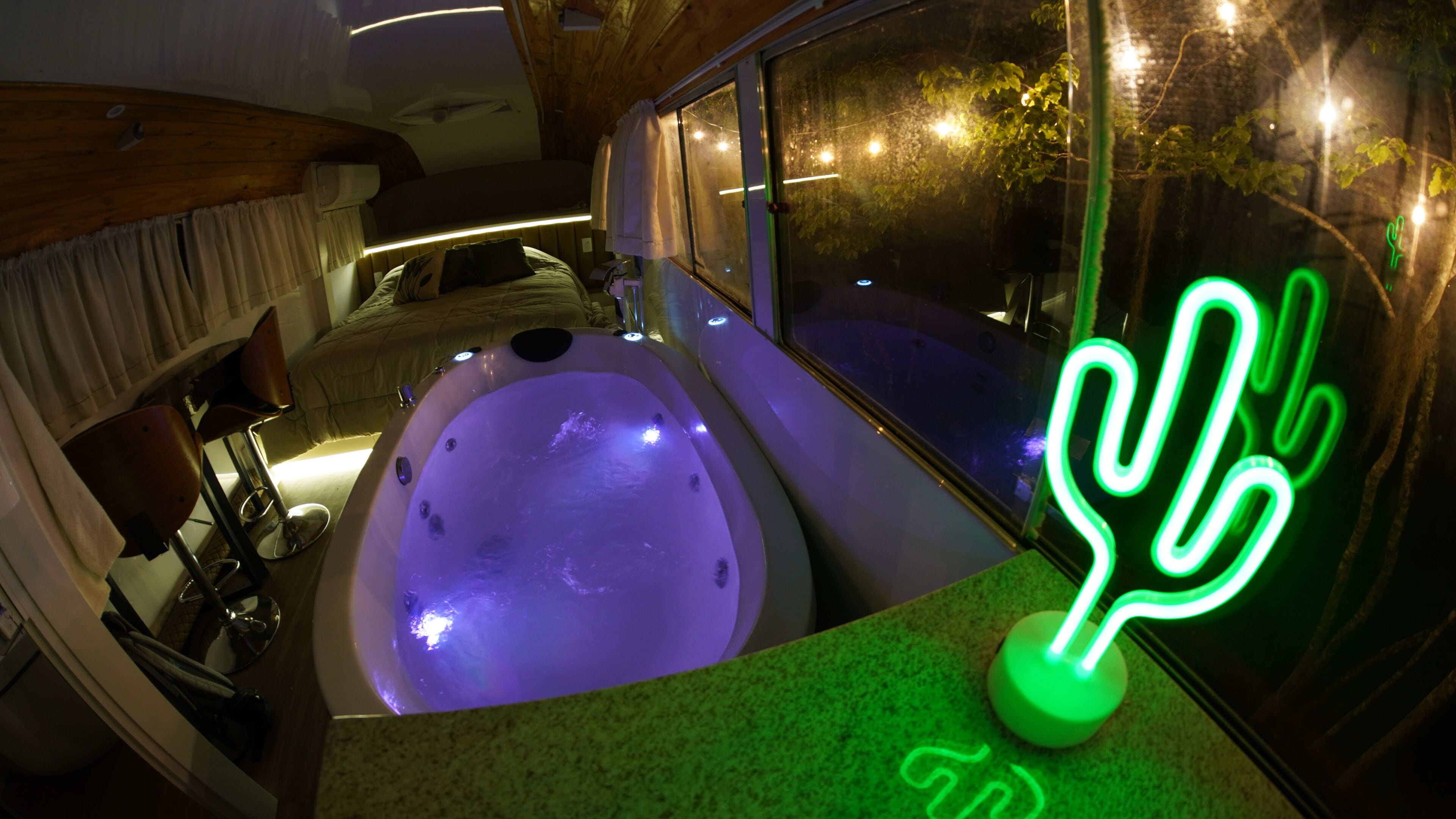 A room with a jacuzzi, a green neon cactus sign, and a window with curtains.