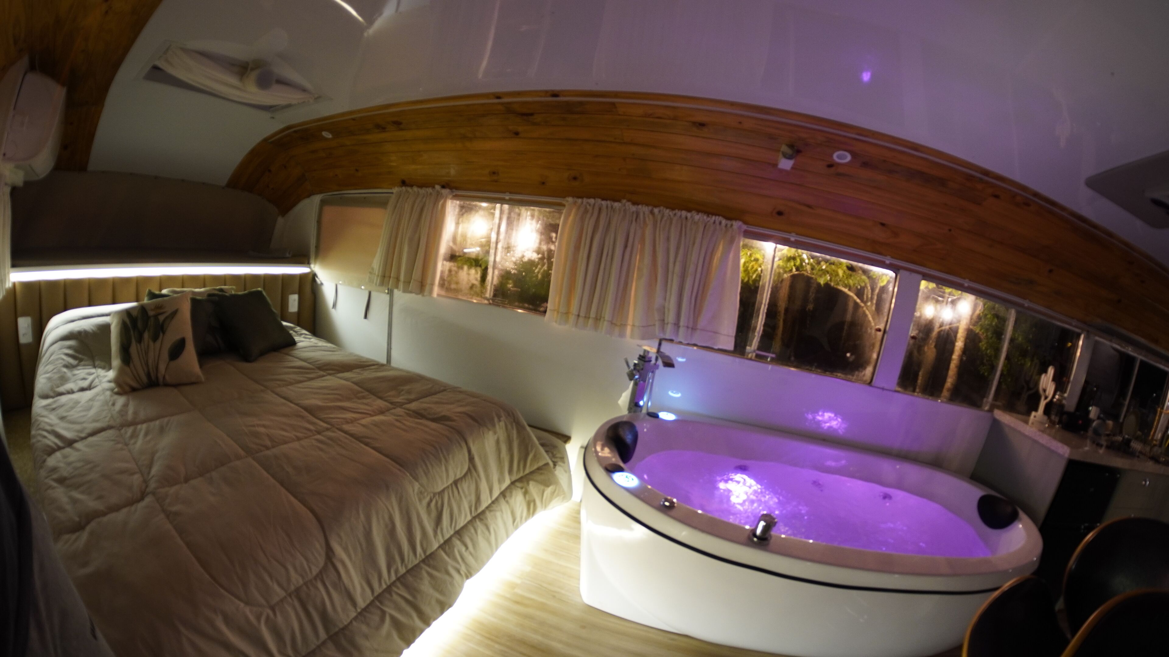 Quarto com cama, janela com cortinas e banheira com iluminação roxa.