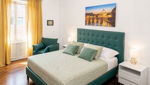 Quarto com uma cama grande, uma mesinha de cabeceira, uma luminária, uma cadeira, uma janela com cortinas e um quadro na parede.