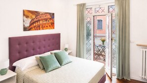 Quarto com uma cama grande, cabeceira, mesa de cabeceira, luminária, pintura do Coliseu e vista para área externa com mesa e cadeiras.