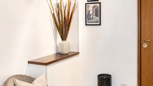 Quarto com piso de madeira, uma cadeira com almofada, uma pequena estante com uma planta, uma lixeira e uma porta.