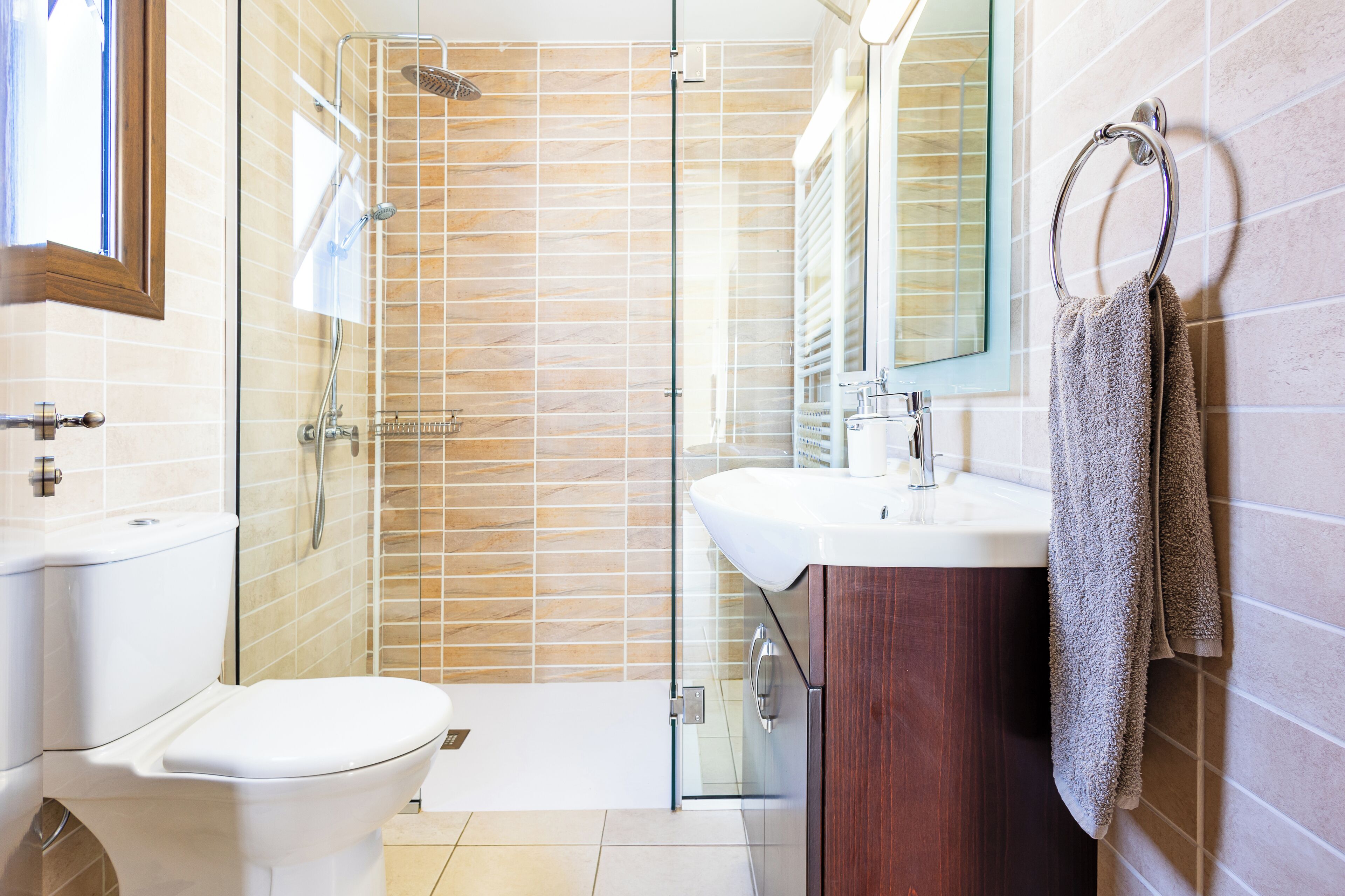 Et moderne badeværelse med brusekabine i glas, et hvidt toilet, et trækommode med spejl og en bøjlestang til håndklæder.