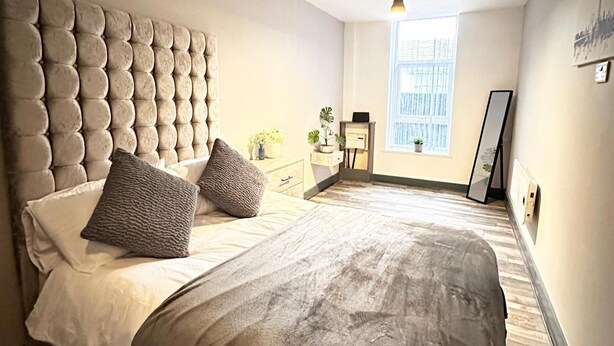 A bedroom with a tufted headboard, a bed with two pillows, a bedside table with a lamp, a window with blinds, and a mirror on the wall. - Oak Suite - 2 Bed - Sleeps 4 - Central Blackpool (Blackpool)