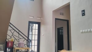 Un'ampia stanza con soffitto alto, pavimento in legno, un tavolo da pranzo con sedie, una scala e una porta che dà su un'altra zona.