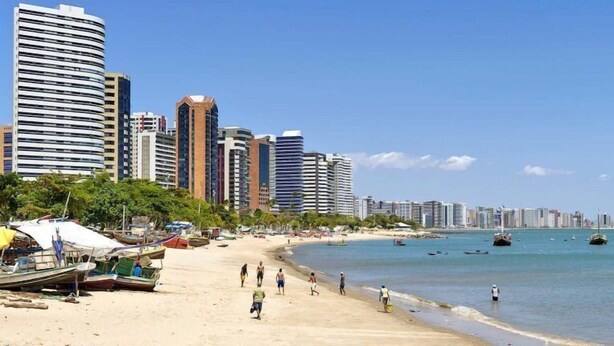 Una spiaggia con una fila di edifici alti sullo sfondo, una battigia sabbiosa e diverse imbarcazioni in acqua.