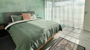 Ein Schlafzimmer mit einem großen Bett, einem Nachttisch, einer Lampe und Blick ins Freie durch transparente Vorhänge.