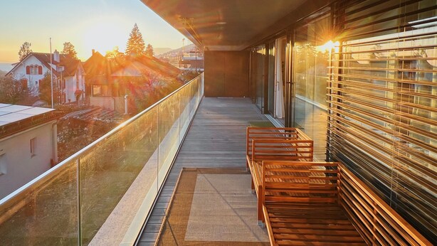 Ein Balkon mit Blick auf Häuser und Bäume, mit hölzernen Bänken und einem Glasgeländer.