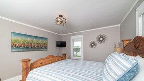 A bedroom with a wooden headboard, a bed with striped bedding, a television on the wall, and a window with a view of greenery.