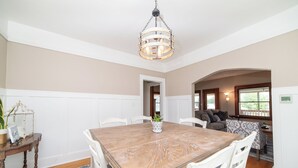 A dining room with a wooden table, white chairs, and a chandelier. There is a plant on the table and a small table with a plant and a sign on the left.