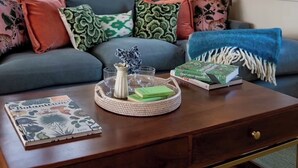 A cozy living room with a grey sofa, colorful cushions, a wooden coffee table with a botanical book, a framed picture of a milk carton, and a window with a view of trees. - Large detached farmhouse in quiet Kent countryside , close to London train line (Edenbridge)