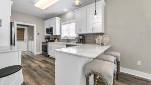 A modern kitchen with white cabinetry, a central island, pendant lighting, and a wooden floor. - Under the Oaks Cottage in Blue Mountain Beach (Santa Rosa Beach)
