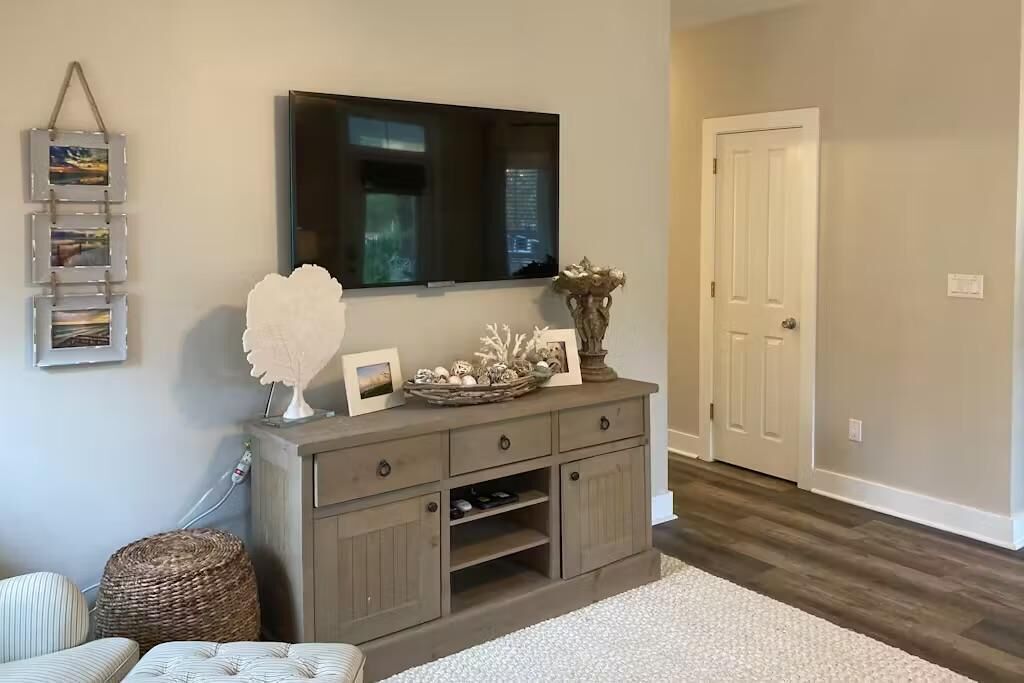 Una sala de estar con un televisor montado en la pared, un mueble de madera con objetos decorativos, una cesta de mimbre y una puerta al fondo.