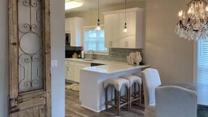 A modern kitchen with white cabinets, a central island with stools, pendant lighting, and a chandelier. - Under the Oaks Cottage in Blue Mountain Beach (Santa Rosa Beach)