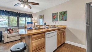A compact kitchen with wooden cabinets, a built-in dishwasher, a stainless steel refrigerator, and a ceiling fan. There are two windows with blinds, a small seating area with a sofa and a chair, and artwork on the walls. - Hidden Beach Villas 122 - A Sunset Paradise (Santa Rosa Beach)