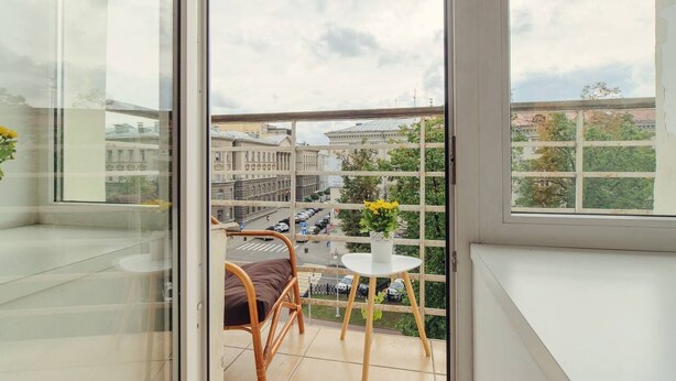 A balcony with a wicker chair and a small table, overlooking a cityscape with buildings and a road.