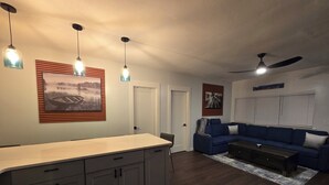 A modern living room with a dark wood floor, a blue sofa, a white coffee table, and a kitchen area with a large cabinet and pendant lights.
