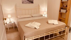 Una cama con ropa de cama cuidada, con cubrecolcha acolchado, dos almohadas y toallas dobladas sobre una estructura de metal blanco. Se observa una mesita de noche con una lámpara, un cuadro en la pared y un estante de madera con cestas.