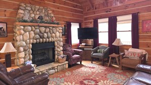 Interior de una cabaña de troncos con chimenea de piedra, muebles de cuero, televisor de pantalla plana y un letrero que dice 'LOVE'.
