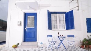 A white building with a blue door and window shutters, a small table with chairs, and a potted plant. - Old Town Traditional House (Paros)
