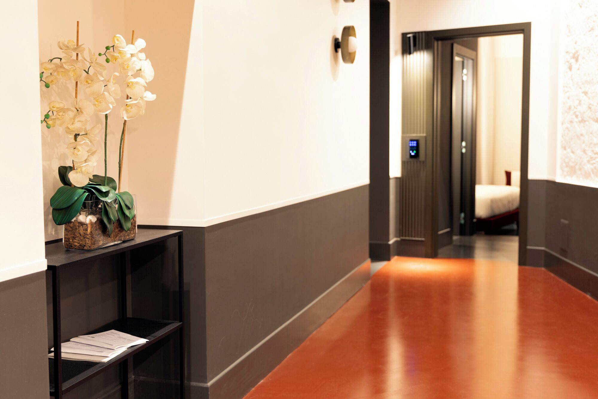 A hallway with a wooden floor, a reception desk with a plant, and a bed visible through a door.