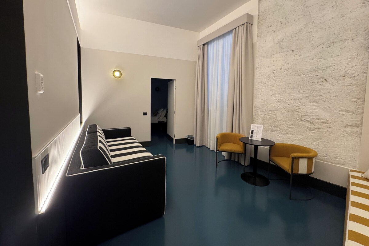 Une chambre d’hôtel moderne, équipée d’un lit, d’un canapé, d’une petite table et d’une chaise. On y trouve une porte donnant sur une autre pièce, ainsi qu’une fenêtre avec des rideaux.