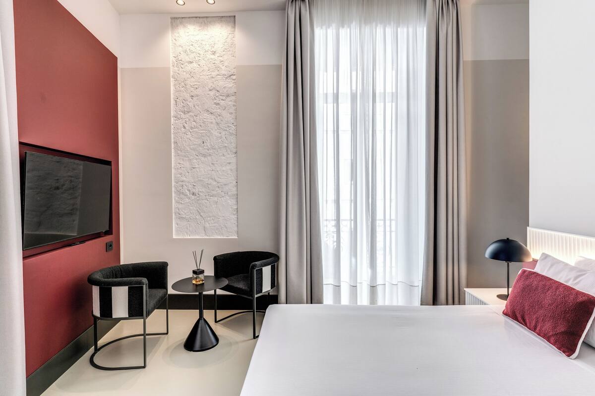 A modern hotel room with a large bed, a flat-screen TV, a small table with two chairs, a wall-mounted artwork, and a view of the outdoors through sheer curtains.