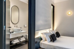 A modern hotel room with a large oval mirror, a white sink, a bedside table, and a bed with white and black pillows. - Adaram Suites (Naples)