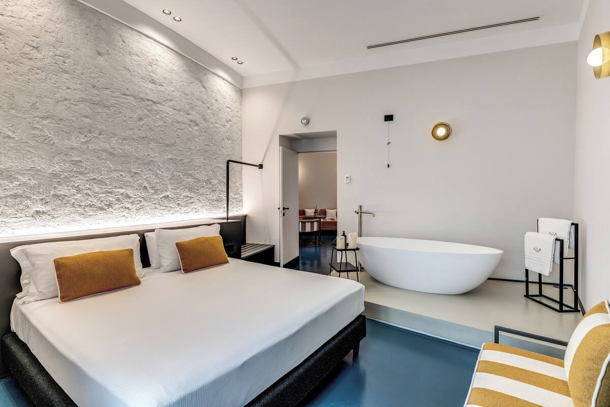 A modern hotel room with a large bed, a bathtub, and a small seating area by the pool.