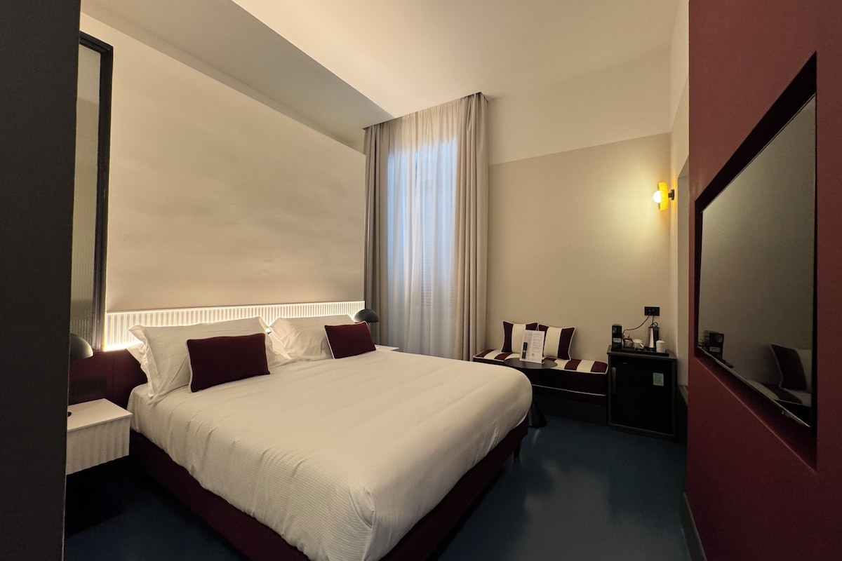 A hotel room with a large bed, bedside tables, a desk, a chair, a mirror, and a window with curtains.