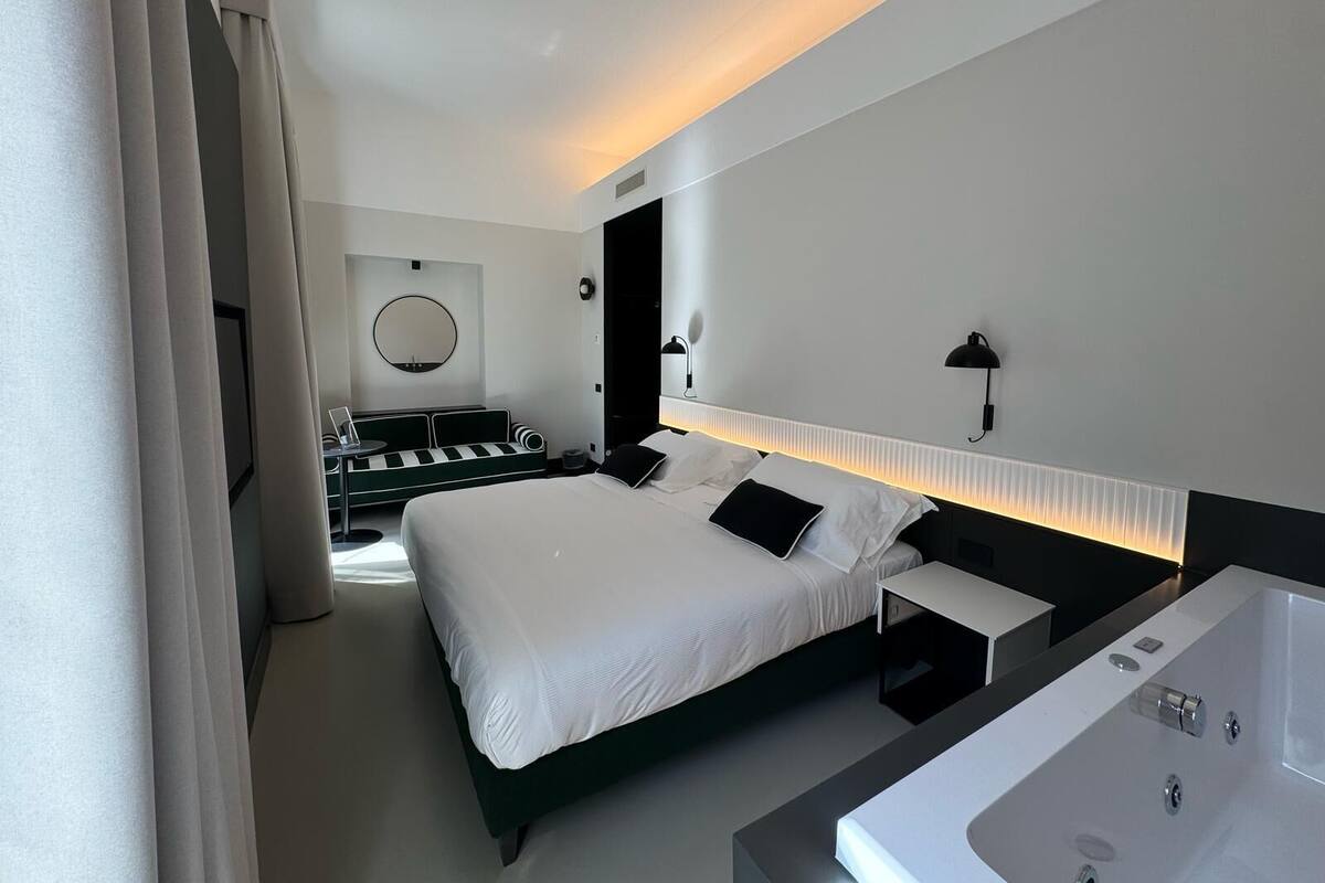 A modern hotel room with a large bed, a bedside table, a built-in bathtub, a round mirror, and a seating area with a sofa and a small table.