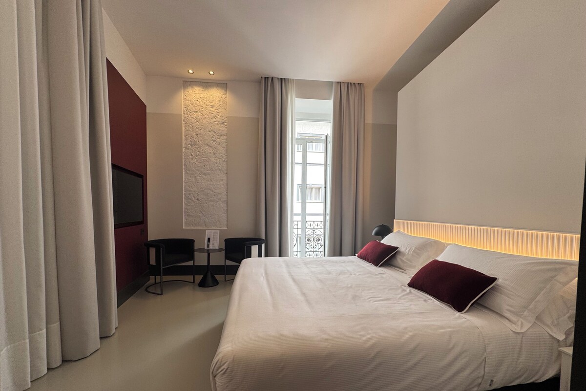 A hotel room with a bed, two chairs, a table, a TV, and a window with curtains.