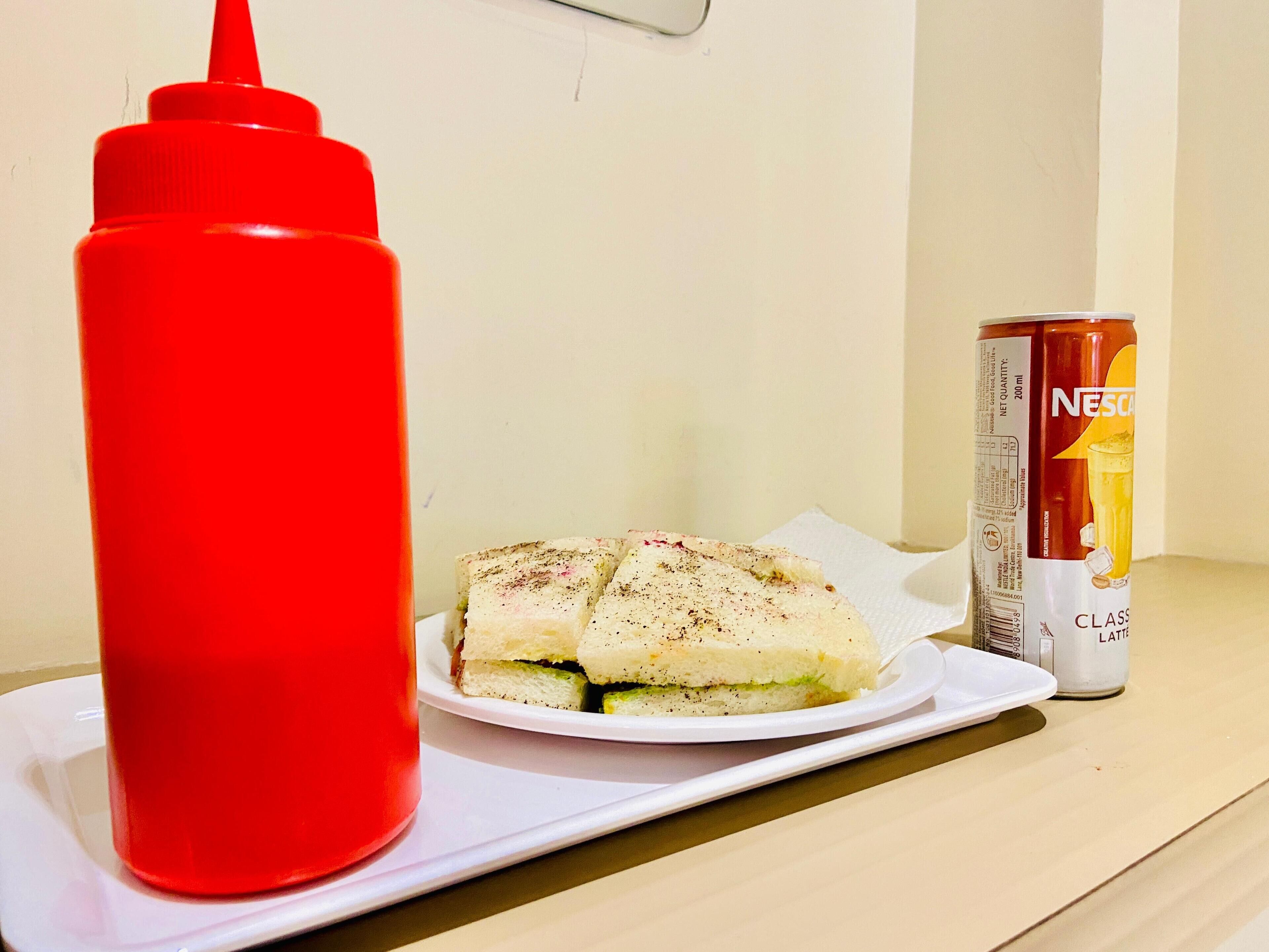 Un sandwich sur une assiette blanche, avec un flacon de sauce rouge et une boîte de Nescafé Class Latte à côté.