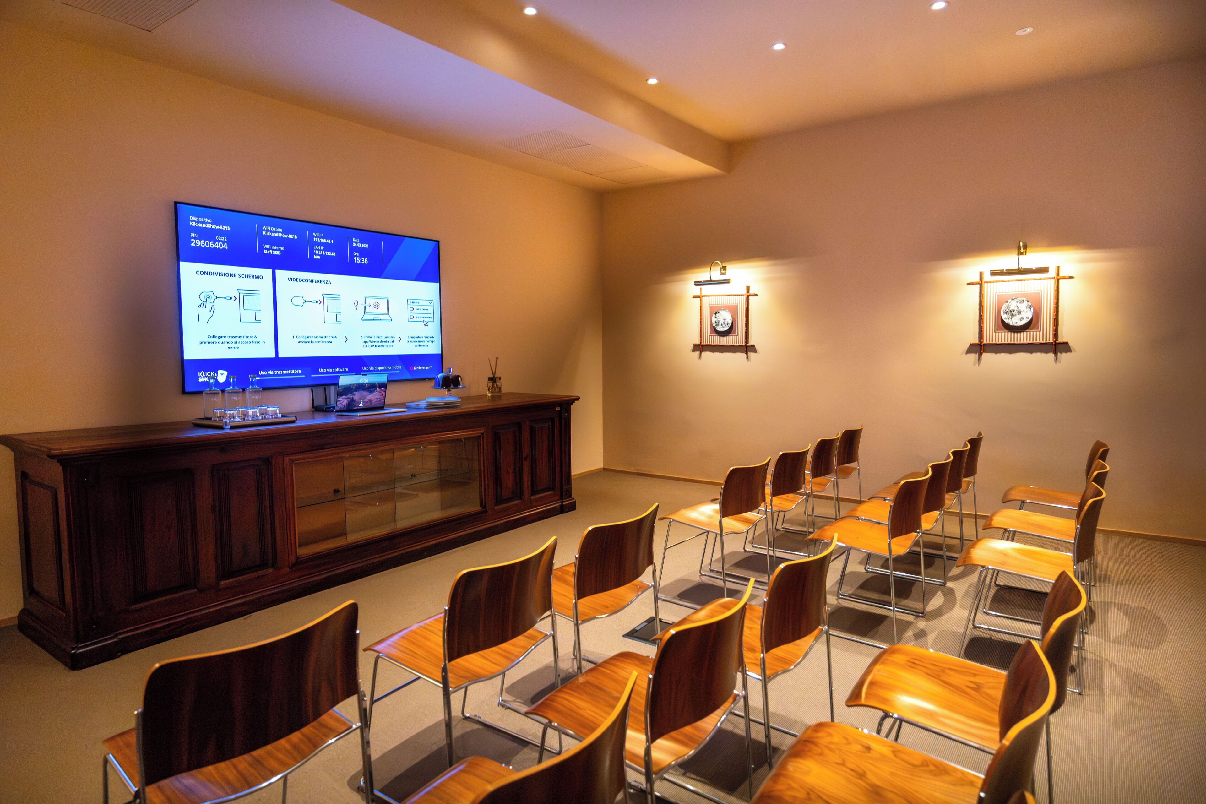 A conference room with a large screen, wooden chairs, a wooden table, and wall-mounted lights.
