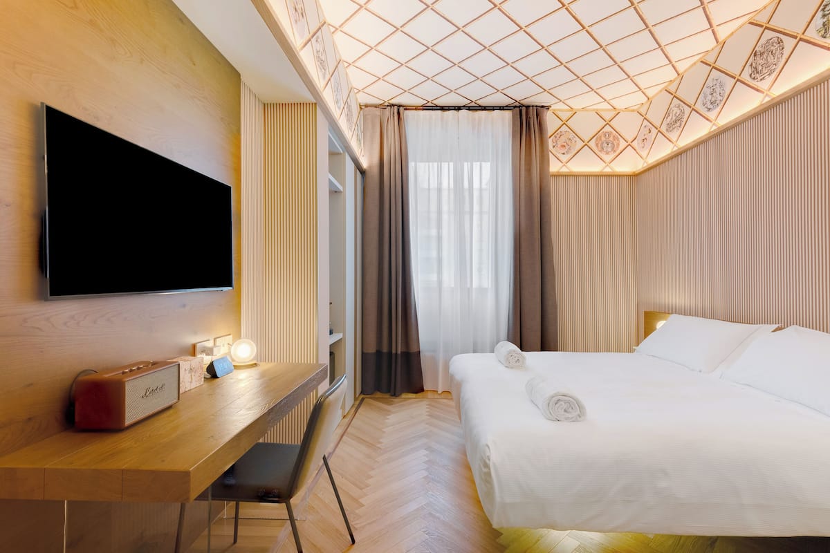 Une chambre d’hôtel comprenant un lit, un bureau avec un téléviseur à écran plat, un haut-parleur et une fenêtre avec des rideaux.