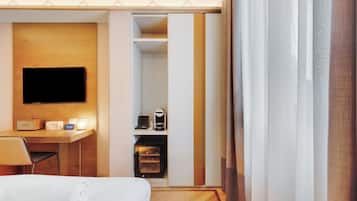 A hotel room with a bed, a desk with a flat-screen TV, a coffee machine, and a minibar.