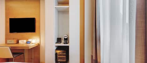 A hotel room with a bed, a desk with a flat-screen TV, a coffee machine, and a minibar.